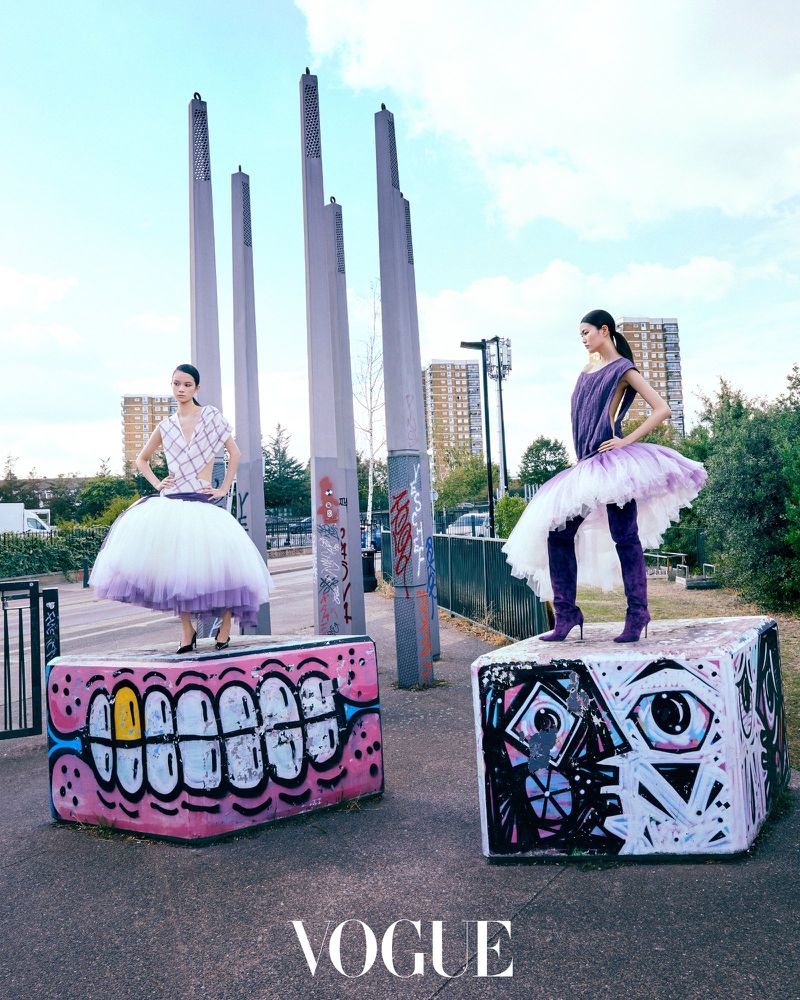 Two models standing on graffiti-painted blocks outdoors wearing voluminous purple-and-white dresses with tall boots