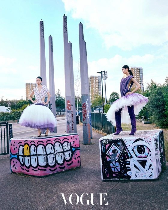 Two models standing on graffiti-painted blocks outdoors wearing voluminous purple-and-white dresses with tall boots
