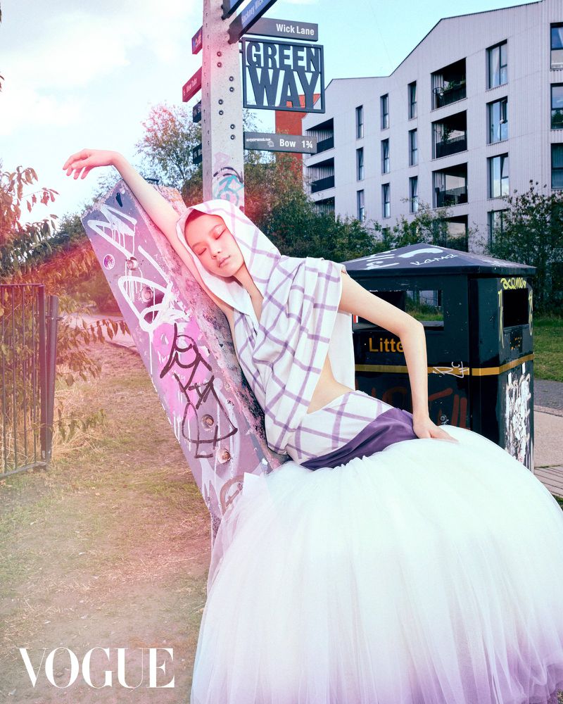 A female model wearing a white hooded top with purple plaid and a voluminous white tulle skirt leaning against a graffiti-covered post outdoors.