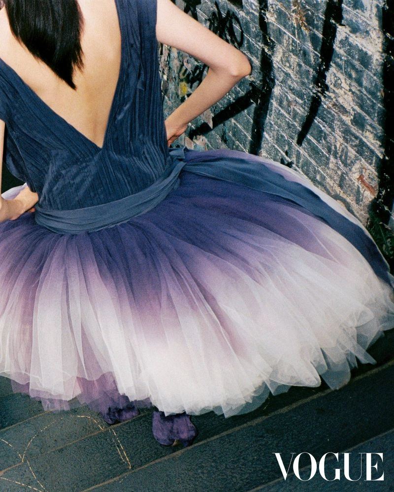 Model seen from behind wearing a dark blue top and voluminous ombre tulle skirt standing against a textured brick wall.