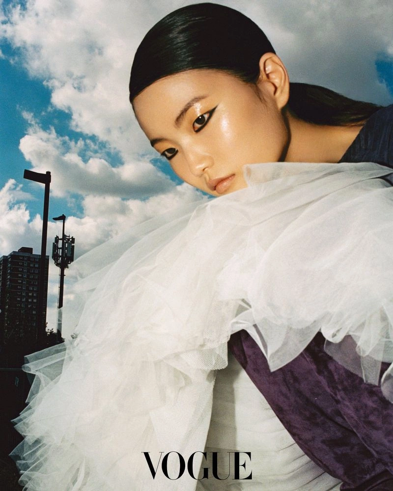 Model with slicked hair and dramatic eyeliner wearing white tulle against a cloudy blue sky with distant buildings
