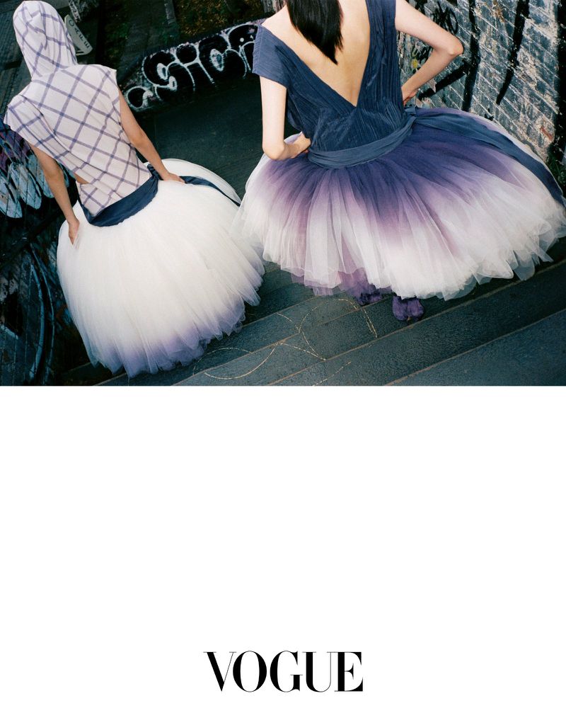 Two models from behind wearing voluminous tulle skirts descending stone stairs with graffiti wall background.