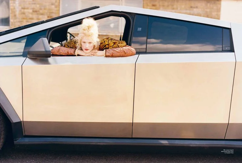 Model with blonde updo resting arms on the window frame of a silver futuristic truck