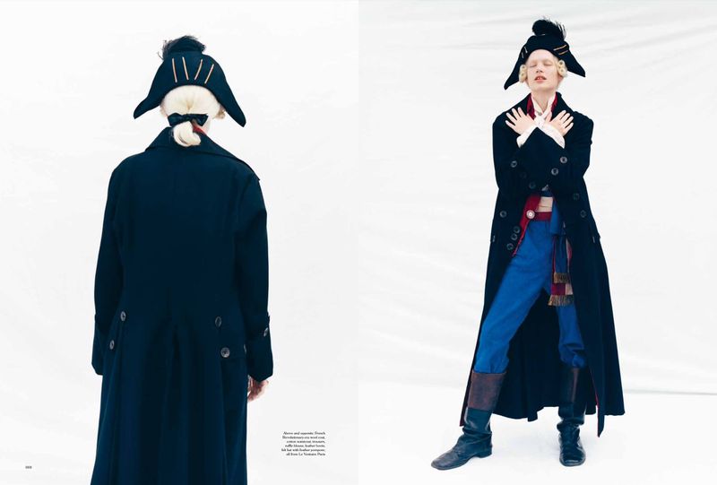Model in historical-inspired black coat, blue trousers, and tricorne hat standing against a plain white background.