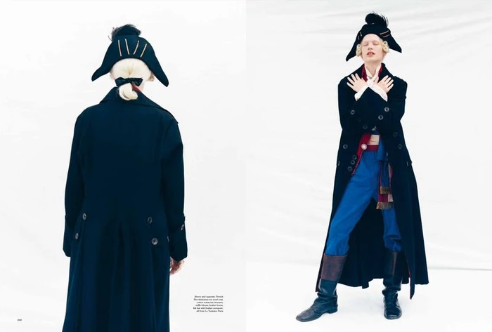 Model in historical-inspired black coat, blue trousers, and tricorne hat standing against a plain white background.