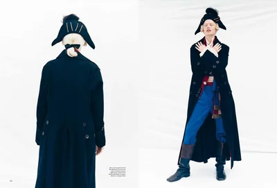 Model in historical-inspired black coat, blue trousers, and tricorne hat standing against a plain white background.