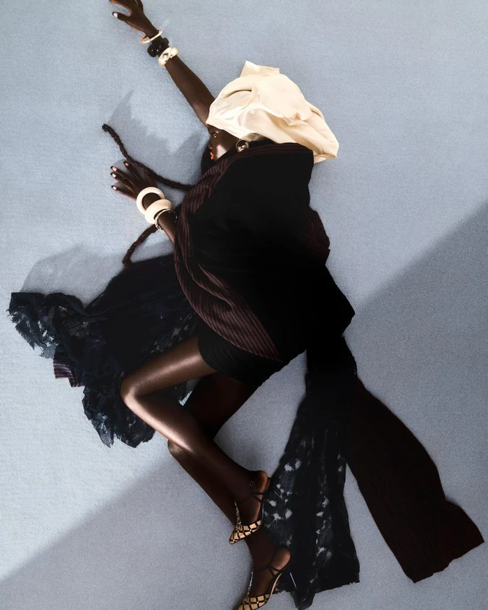 Model Achol Kuir lying on a light grey studio floor wearing dark clothing and a cream headwrap