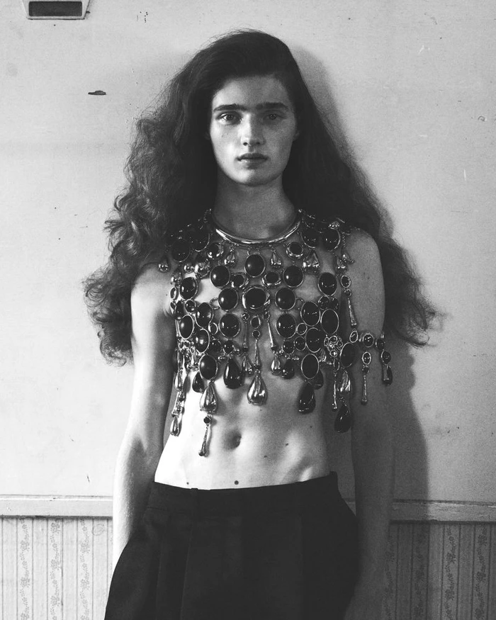 Model with long dark wavy hair wearing an elaborate jeweled collar and dark trousers against a textured wall.