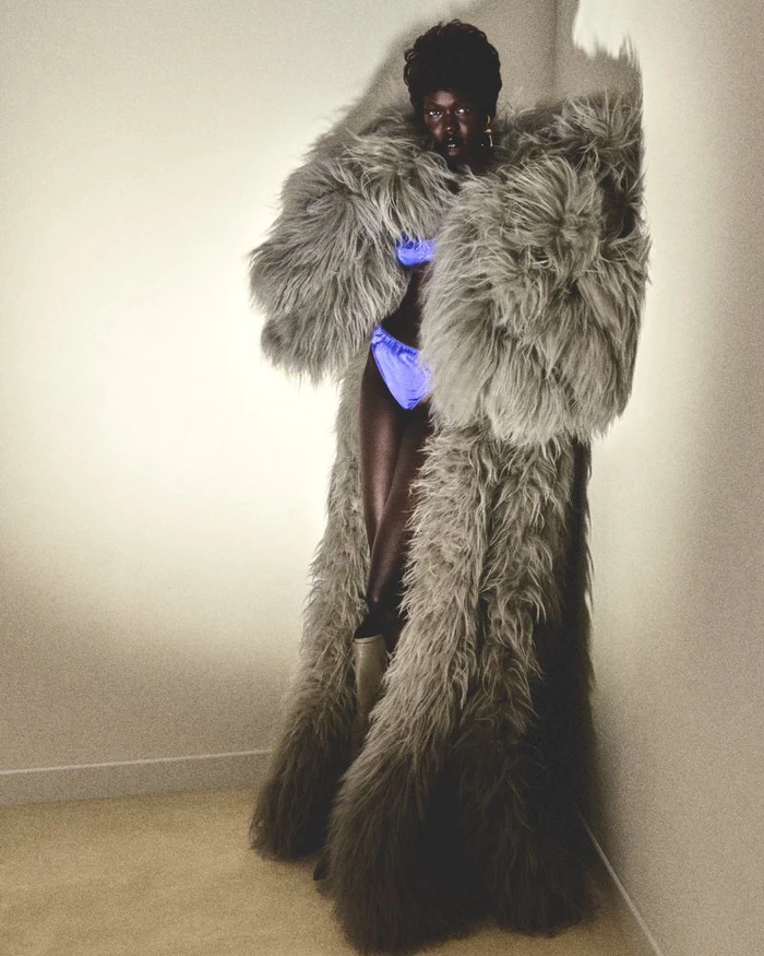 Model Achol Kuir wearing a massive grey shaggy fur coat and blue satin underwear standing against a light wall