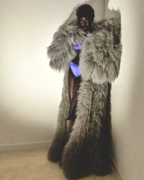 Model Achol Kuir wearing a massive grey shaggy fur coat and blue satin underwear standing against a light wall