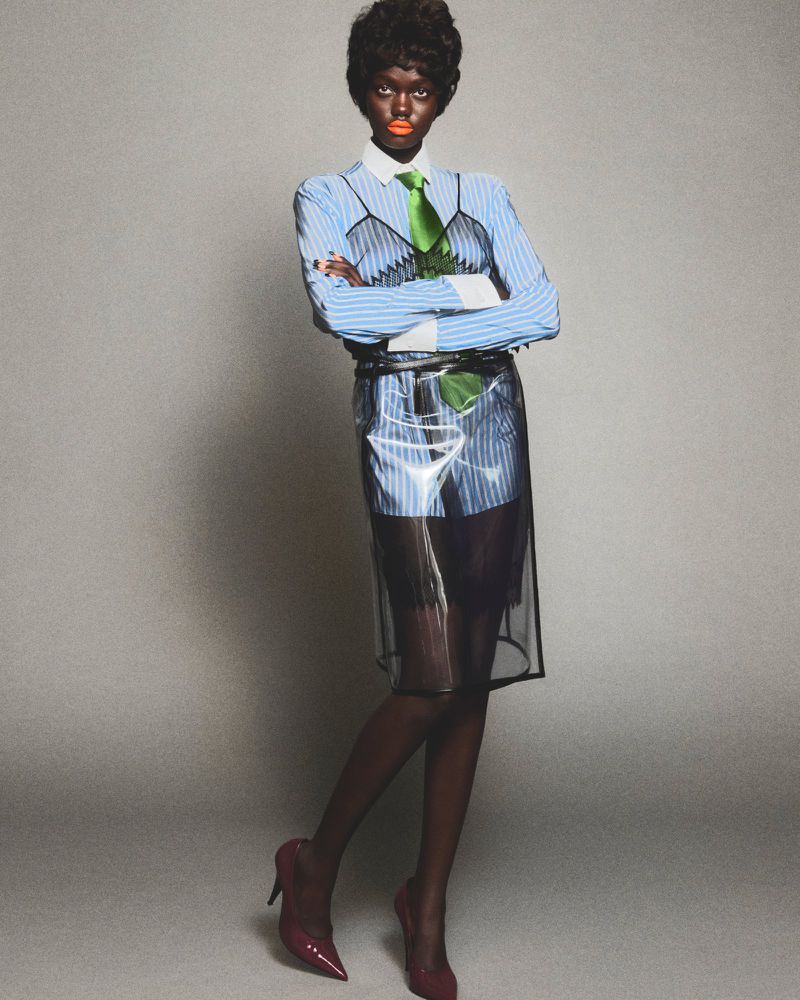 Model Achol Kuir in a blue striped shirt, green tie and transparent plastic skirt against a grey background