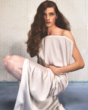 Model with long brown hair wearing a structured white strapless garment crouching indoors.
