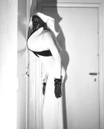 Model Achol Kuir wearing a white bodysuit and headpiece leaning against a doorframe in a monochrome fashion editorial portrait.