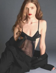 Model with long wavy brown hair wearing a sheer black top and trousers against a grey backdrop for Love Magazine.