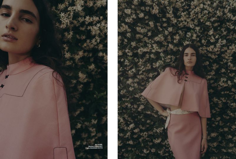 A woman in a pink skirt suit poses against a background of small white flowers in a fashion editorial shot with soft, diffused lighting.