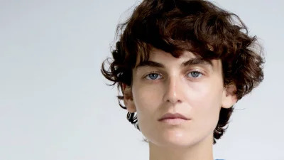 Close-up portrait of a woman with short dark curly hair and blue eyes against a light background
