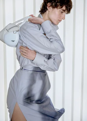Model with short brown hair wearing a light blue shirt and gradient skirt holding a white handbag against vertical slats