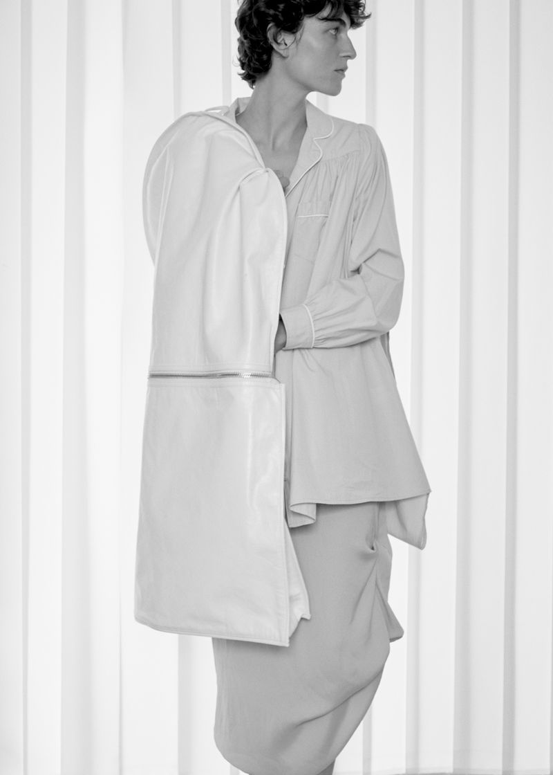 Model in light-colored long-sleeve top holding a garment bag against a vertical slat background