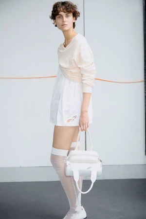 Model with short dark curly hair wearing cream top and white mini dress holding a white handbag against plain wall