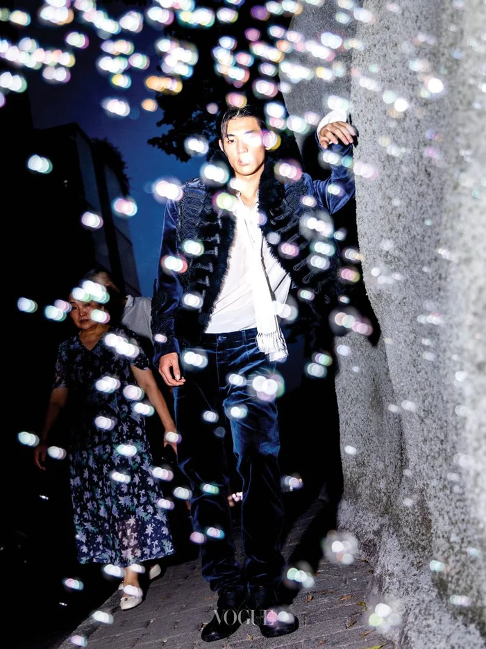 Man in dark velvet jacket standing outdoors at night with blurred bokeh lights floating in the foreground