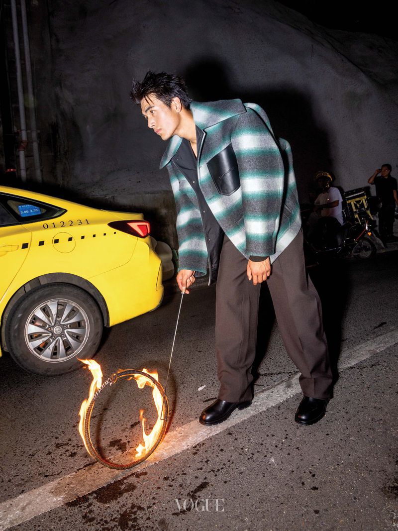 Man in a green and white striped jacket holding a flaming metal hoop beside a yellow taxi on a street at night.