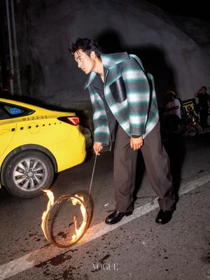 Man in a green and white striped jacket holding a flaming metal hoop beside a yellow taxi on a street at night.