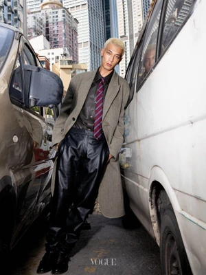 Man with bleached blond hair wearing a grey coat and striped tie leaning between two vehicles on a city street
