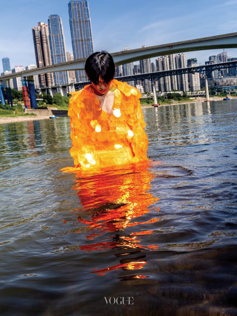 Man wearing a bright orange sequined jacket standing in a river with city skyline and bridges in the background