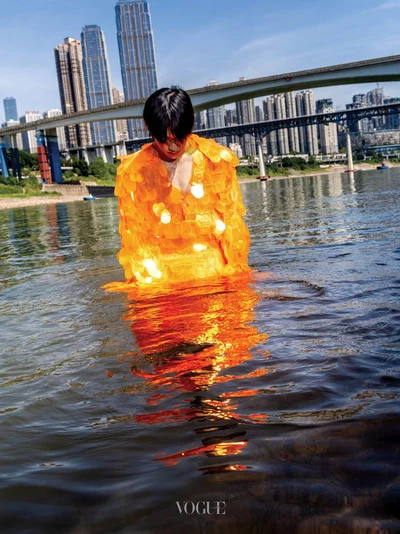 Man wearing a bright orange sequined jacket standing in a river with city skyline and bridges in the background