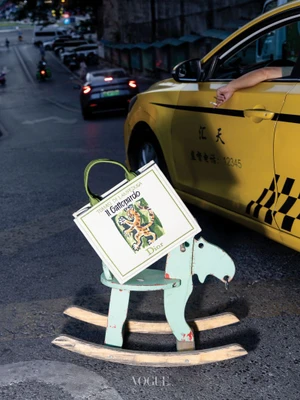 A white tote bag resting on a pale green rocking horse on a street beside a yellow taxi cab.
