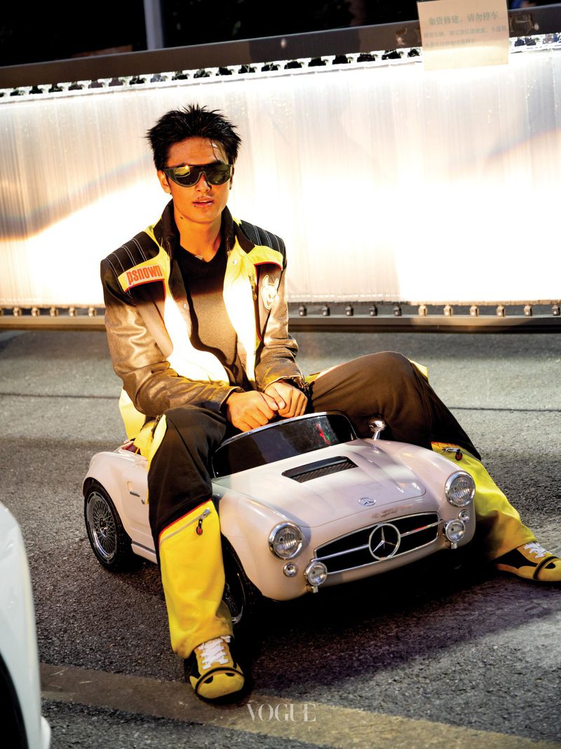 Man wearing sunglasses and a black and yellow jacket straddling a small toy car on a street at night.