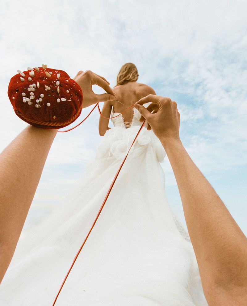 POV shot of hands pulling a red thread from a pincushion toward a bride in a white gown against a pale sky