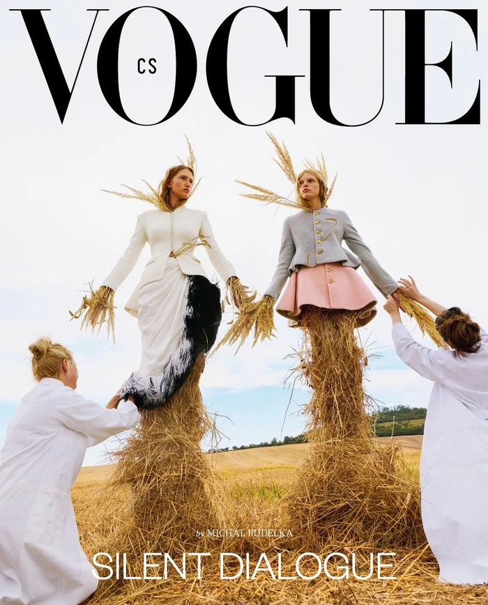 Two female models styled as scarecrows in haystacks in a field, assisted by two figures in white coats.