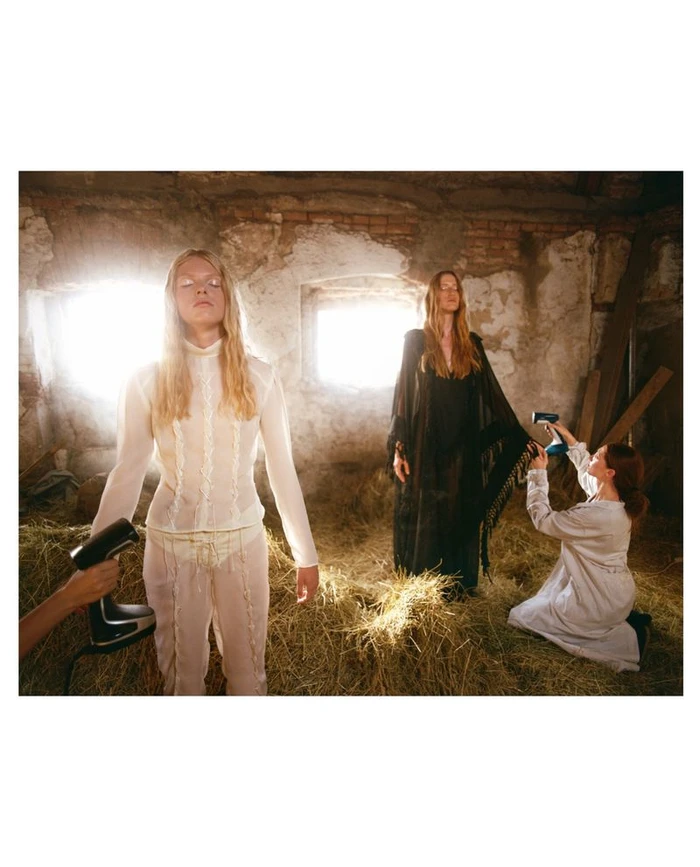 Models in a rustic barn with hay on the floor while crew members use garment steamers on their clothes