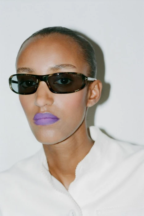 Close-up portrait of Muna Mahamed wearing dark tortoiseshell sunglasses and vibrant purple lipstick against a plain white wall.