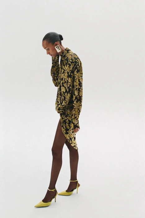 Model Muna Mahamed in a black and gold patterned dress with yellow heels against a white studio background.