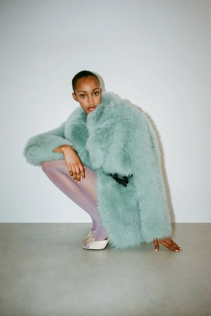 Model Muna Mahamed crouching in a fluffy mint green coat and purple tights against a white seamless background.