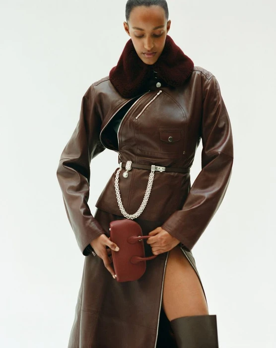 Model Muna Mahamed wearing a brown leather jacket and skirt holding a handbag against a white background