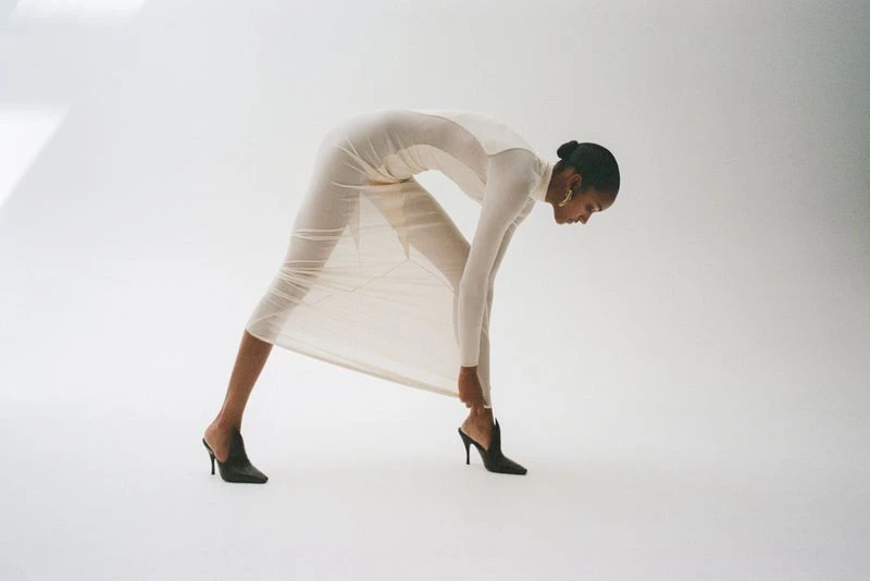Model Muna Mahamed bending forward in a cream dress and black heels against a plain white studio background.