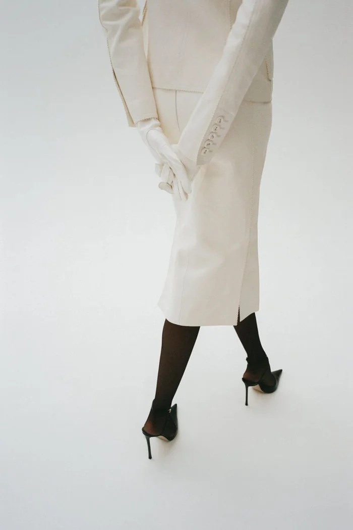 Rear view of a model in a cream skirt suit, white gloves, black tights and heels against a white background