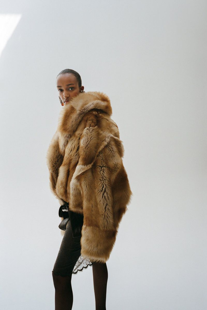 Model wearing a voluminous fluffy tan fur jacket over dark clothing against a bright white studio backdrop.