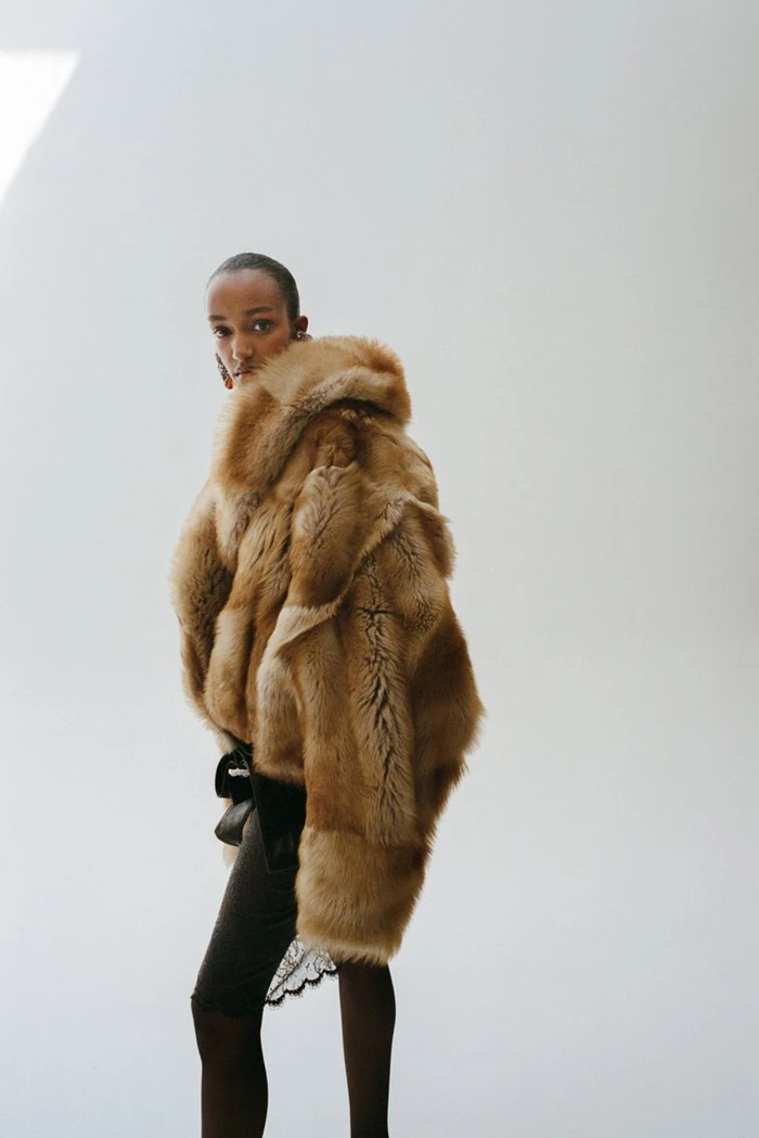 Model wearing a voluminous fluffy tan fur jacket over dark clothing against a bright white studio backdrop.