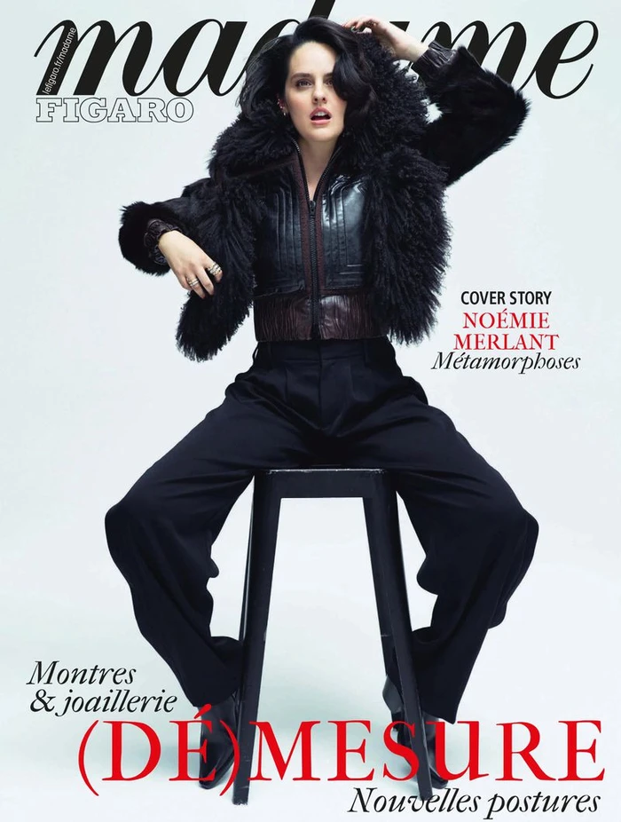 Noemie Merlant sitting on a stool wearing a black furry jacket and dark trousers against a white backdrop for Madame Figaro