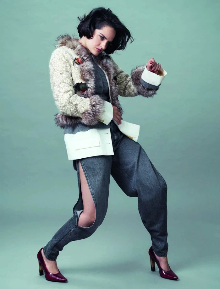 Noemie Merlant in patterned jacket with fur trim and side-slit denim pants posing against a muted green backdrop