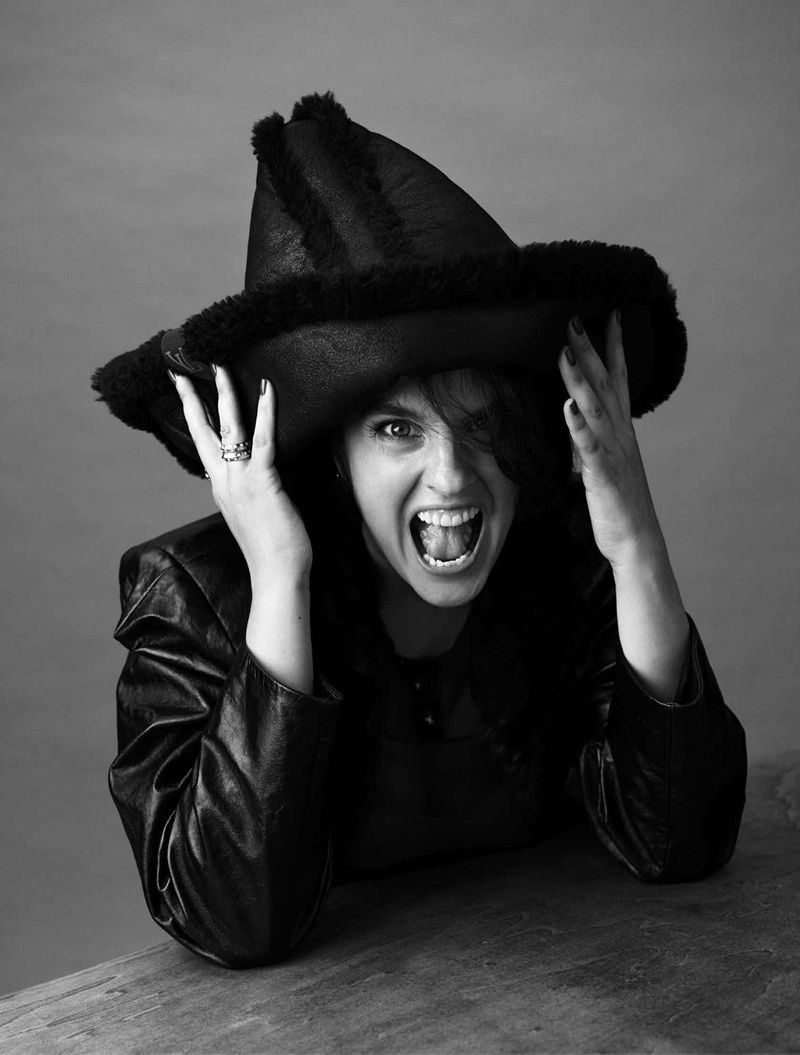 Monochrome portrait of Noémie Merlant wearing a wide-brimmed shearling hat and leather jacket, shouting playfully with hands on brim.