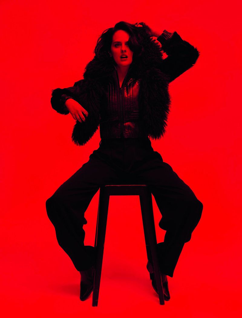 Noemie Merlant seated on a stool wearing dark clothing against a vibrant red background for Madame Figaro