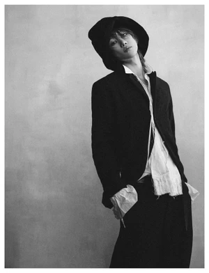 Monochrome fashion portrait of a person in a black jacket and wide-brimmed hat against a textured neutral background