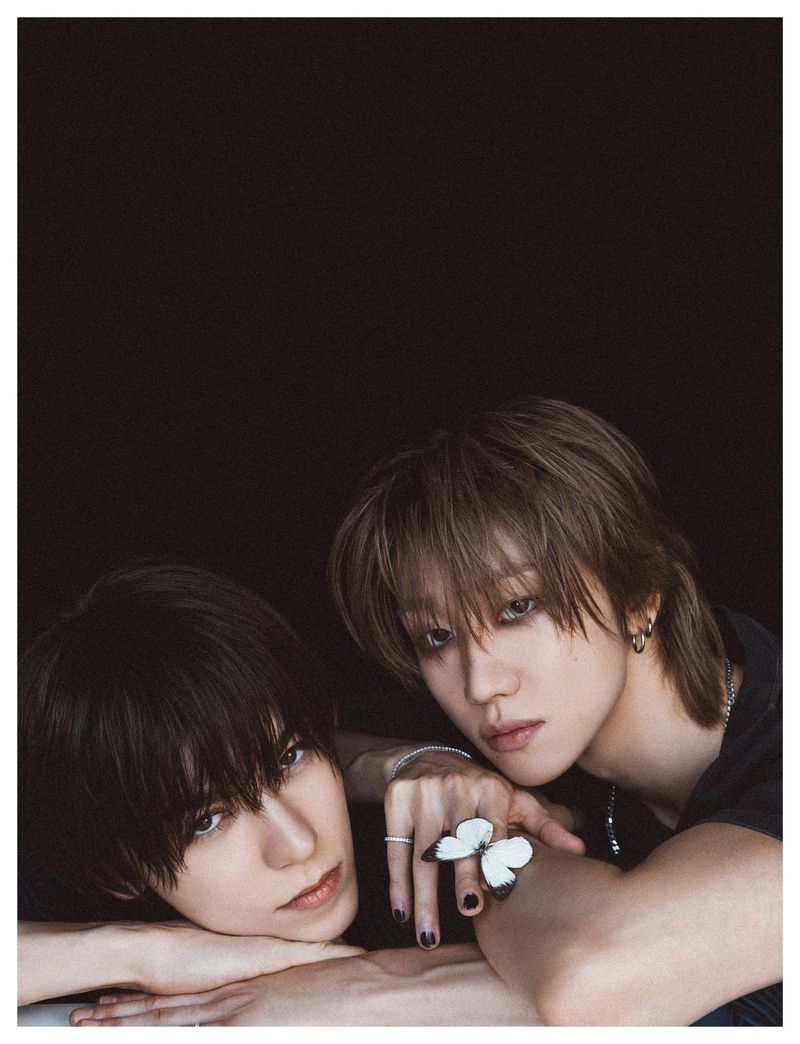 Two young men with dark hair resting their arms against a surface against a dark background, one wearing a butterfly ring
