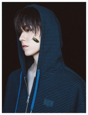 Close-up portrait of a young man wearing a blue striped hoodie with an iridescent beetle resting on his cheek against a dark background