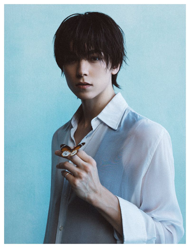 A young man with dark tousled hair wearing a white shirt holding a butterfly on his finger against a light blue background.